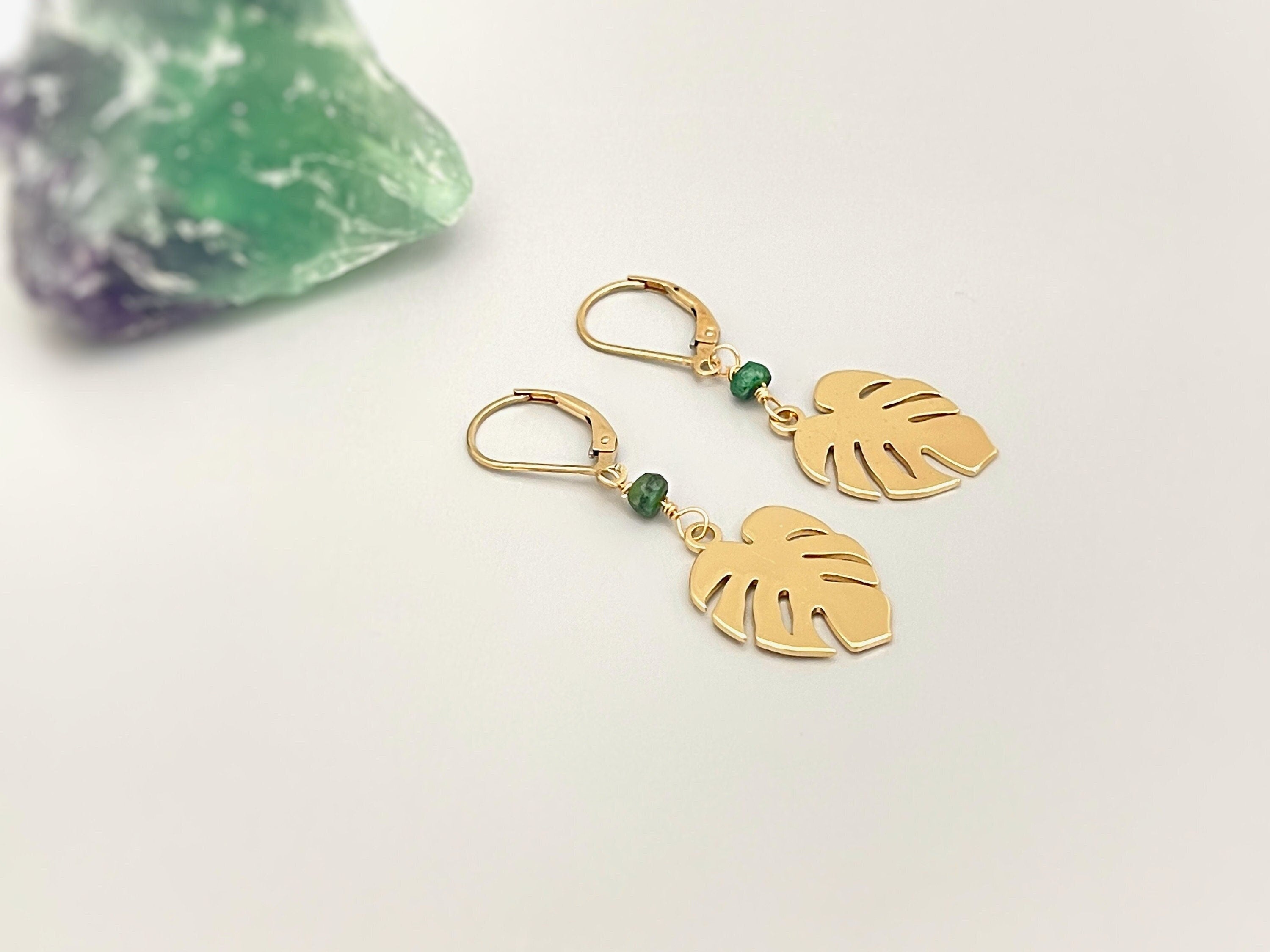 May include: Gold earrings with green gemstone accents and a leaf design. The earrings are dangling and have a hook closure.