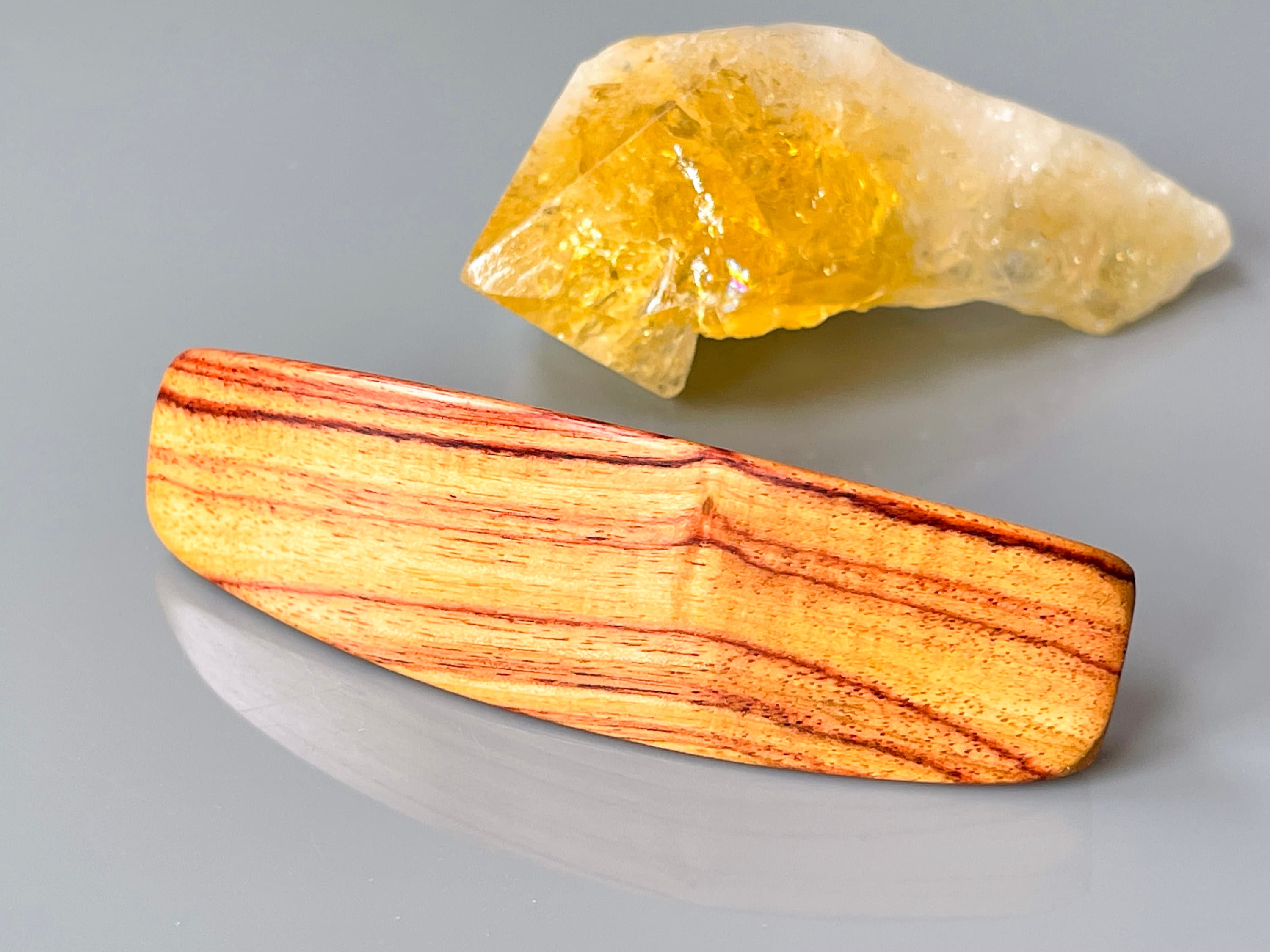 May include: A light brown wooden hair clip with reddish-brown wood grain. The clip is oblong and slightly curved. It is shown next to a yellow-orange crystal cluster.