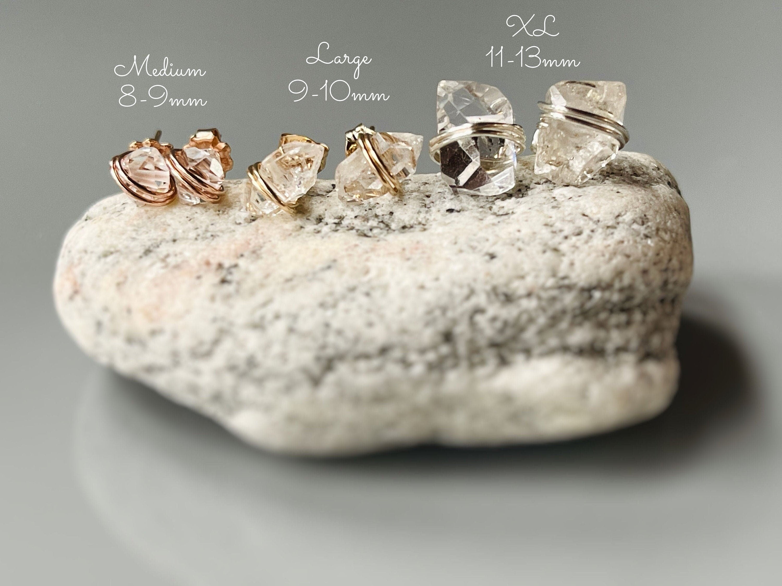 May include: Three pairs of clear quartz crystal earrings with gold wire wrapping. The earrings are arranged in a row from smallest to largest. The smallest earrings are labeled 'Medium 8-9mm', the middle earrings are labeled 'Large 9-10mm', and the largest earrings are labeled 'XL 11-13mm'.