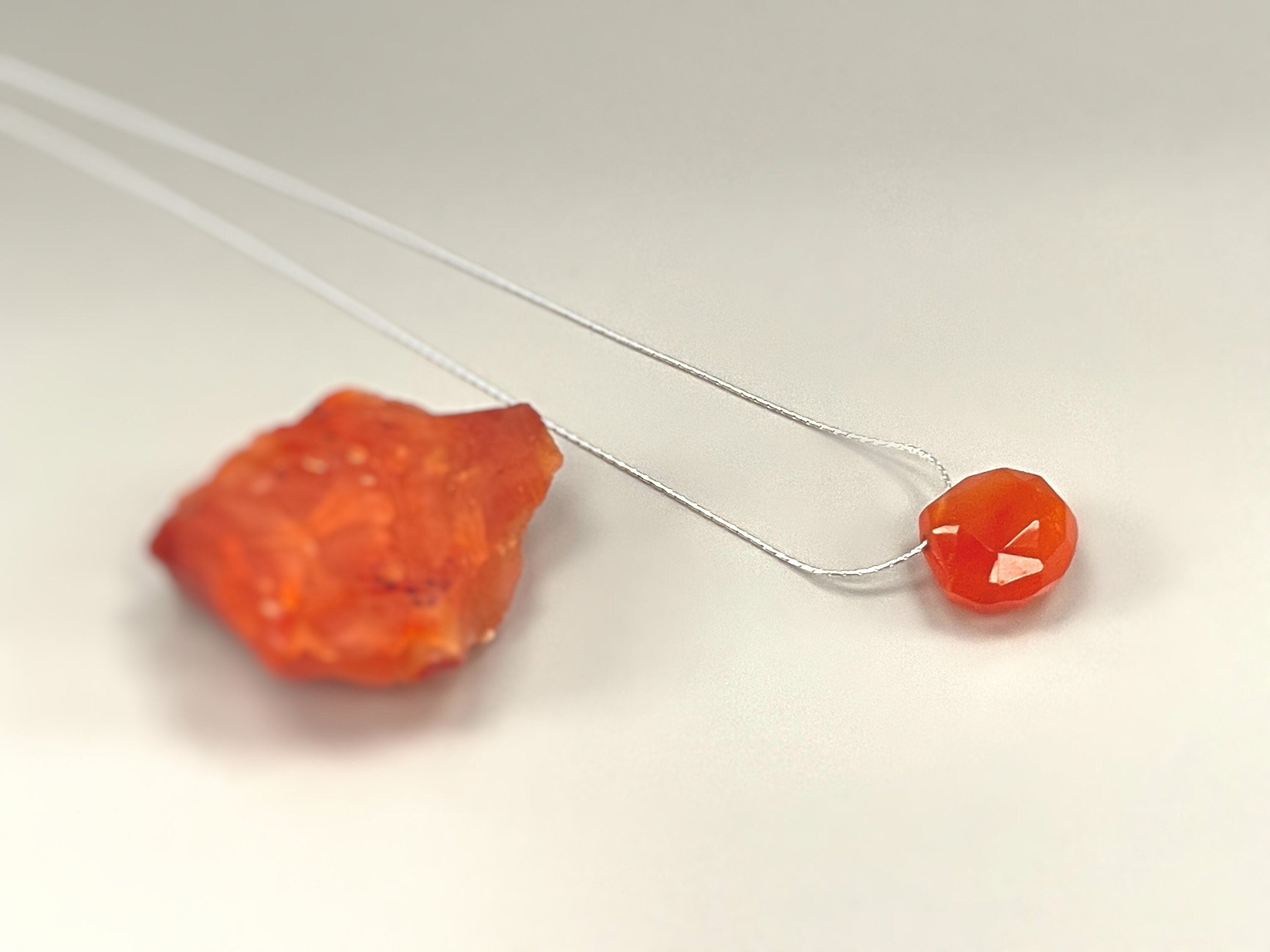 May include: A close-up of a carnelian necklace with a faceted carnelian pendant. The necklace is on a white surface.