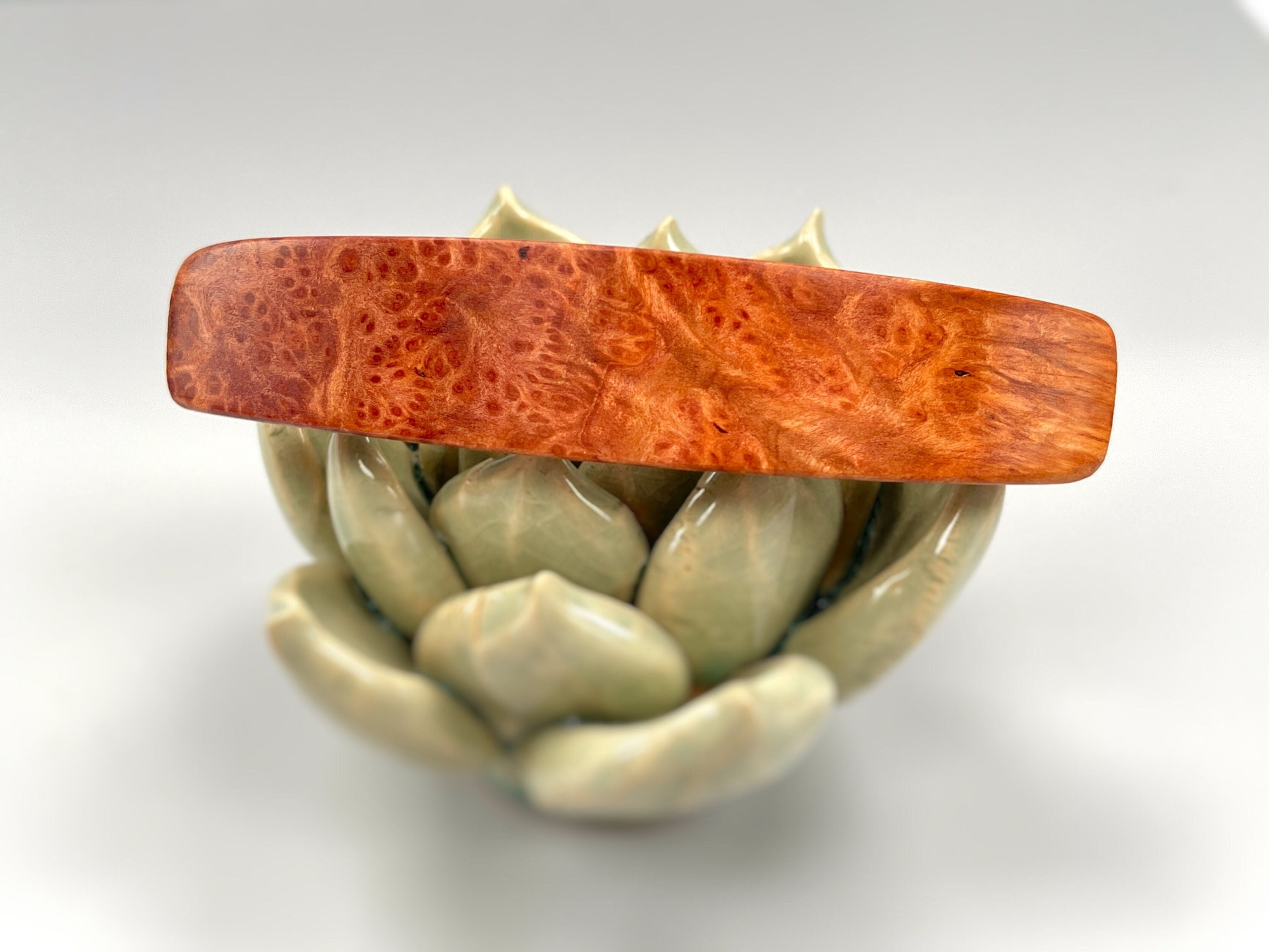 May include: A wooden hair clip with a red and brown wood grain pattern. The clip is shaped like a rectangle and is resting on a green ceramic flower pot.