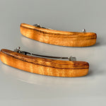 May include: Two wooden hair clips with a silver metal clasp. The clips are curved and have a natural wood grain pattern. The wood is a light brown color with darker brown streaks.