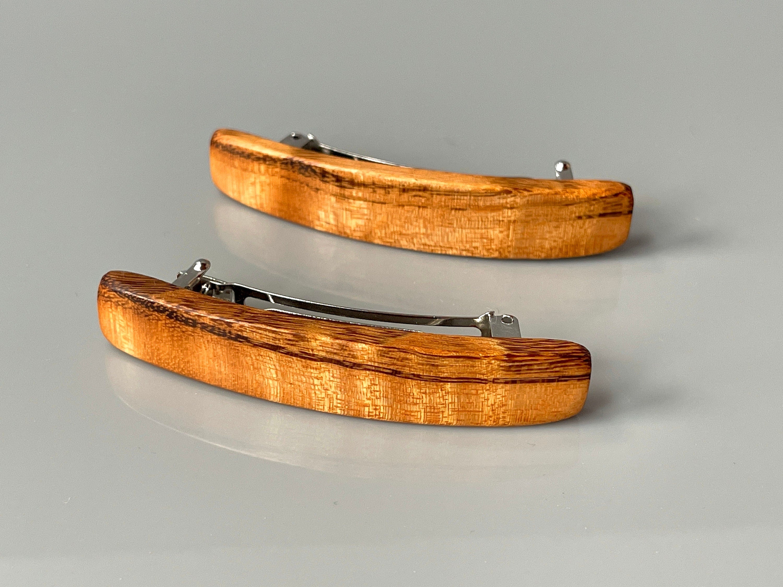 May include: Two wooden hair clips with a silver metal clasp. The clips are curved and have a natural wood grain pattern. The wood is a light brown color with darker brown streaks.