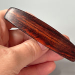 May include: A dark brown wooden hair clip with a smooth, polished finish. The clip has a rectangular shape and is slightly curved. The wood grain is visible and has a rich, dark brown color.