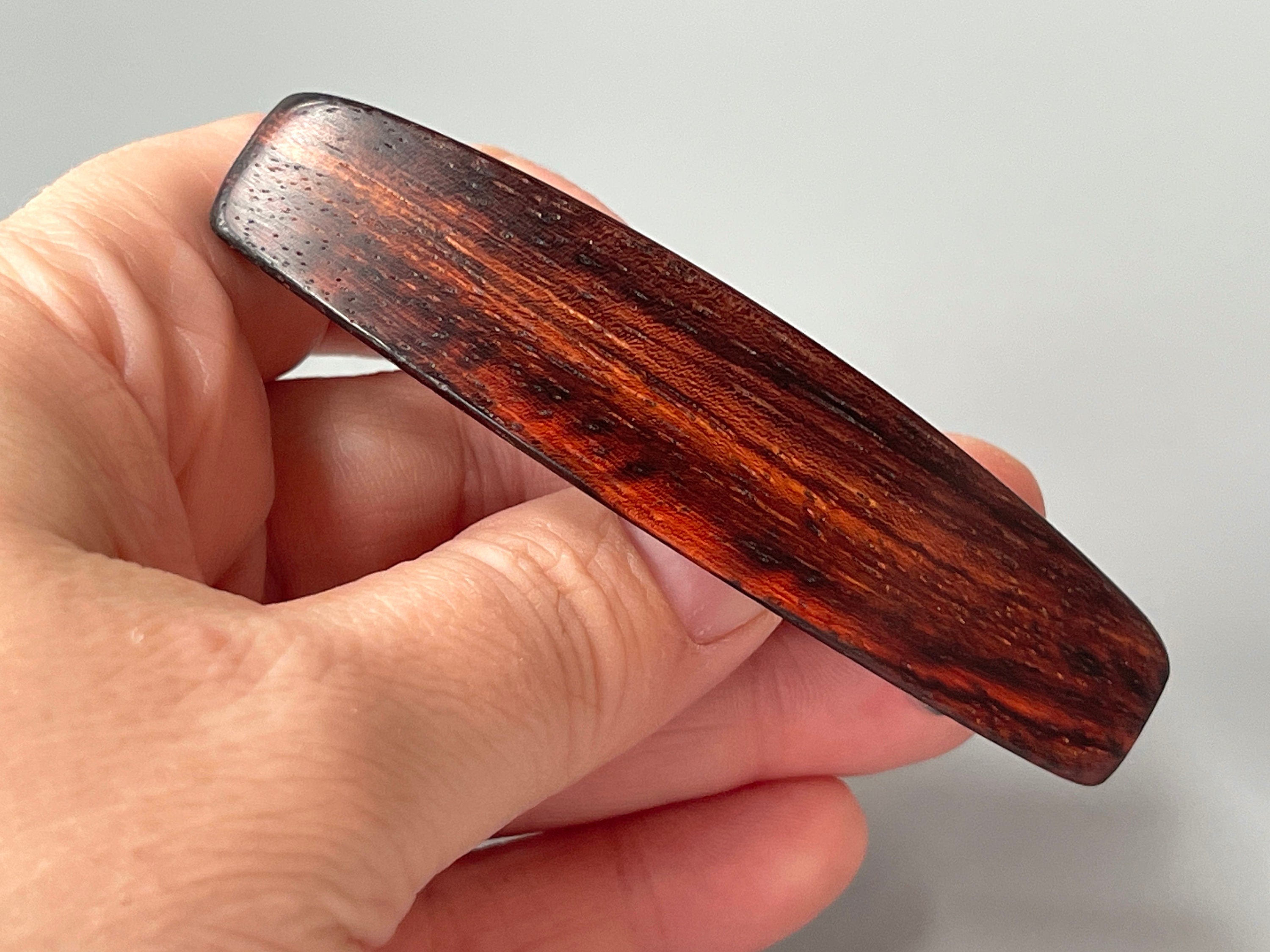 May include: A dark brown wooden hair clip with a smooth, polished finish. The clip has a rectangular shape and is slightly curved. The wood grain is visible and has a rich, dark brown color.
