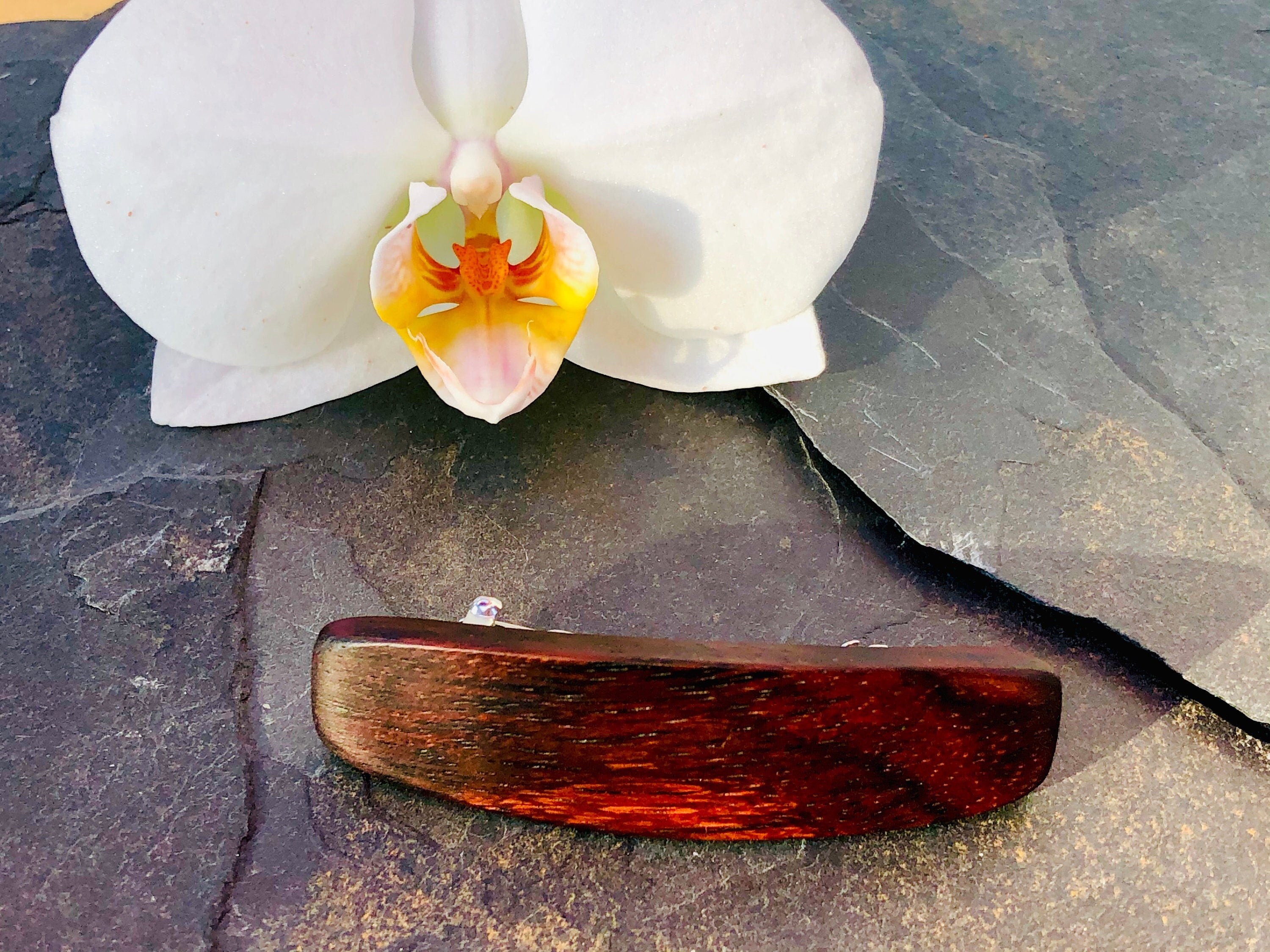 May include: A dark reddish-brown wooden hair clip. The clip is oblong and slightly curved. It has a smooth, polished finish, showing the wood grain. The hair clip is displayed on a dark gray slate surface, next to a white orchid with a yellow center.