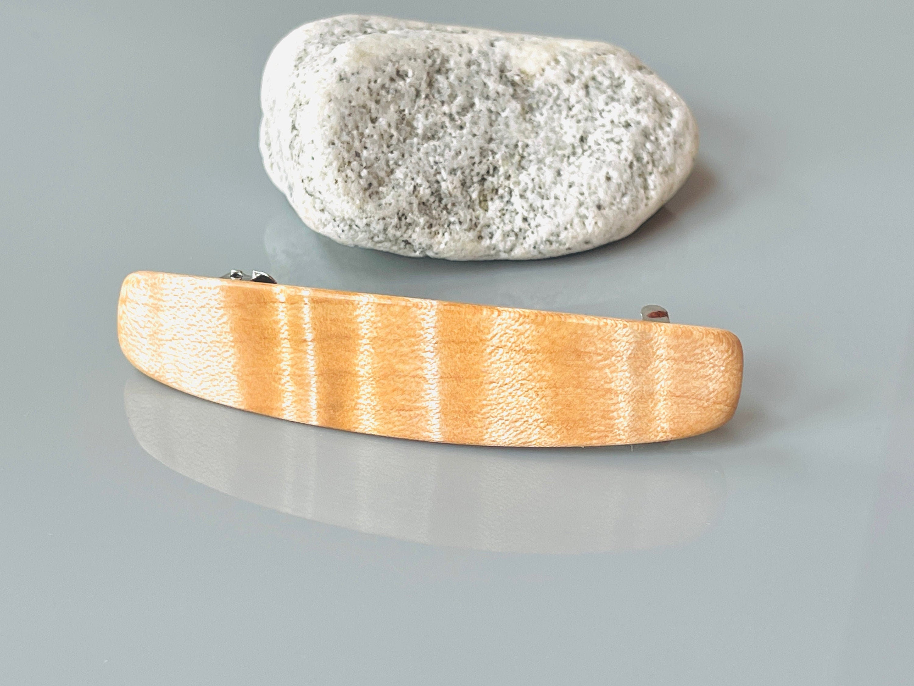 May include: A light-colored wooden hair clip with a subtle striped pattern. The clip is gently curved and features metallic silver-toned clasps on either end. It's showcased against a neutral gray background, next to a light gray stone.