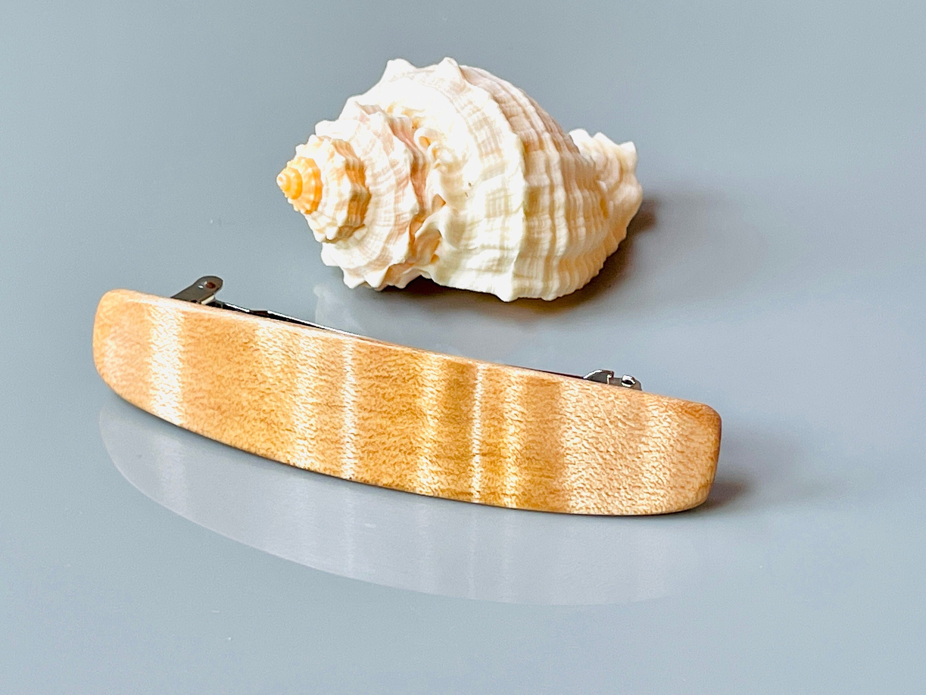 May include: A light brown wooden hair clip with a silver metal clasp. The clip has a smooth, polished finish and a slightly curved shape.