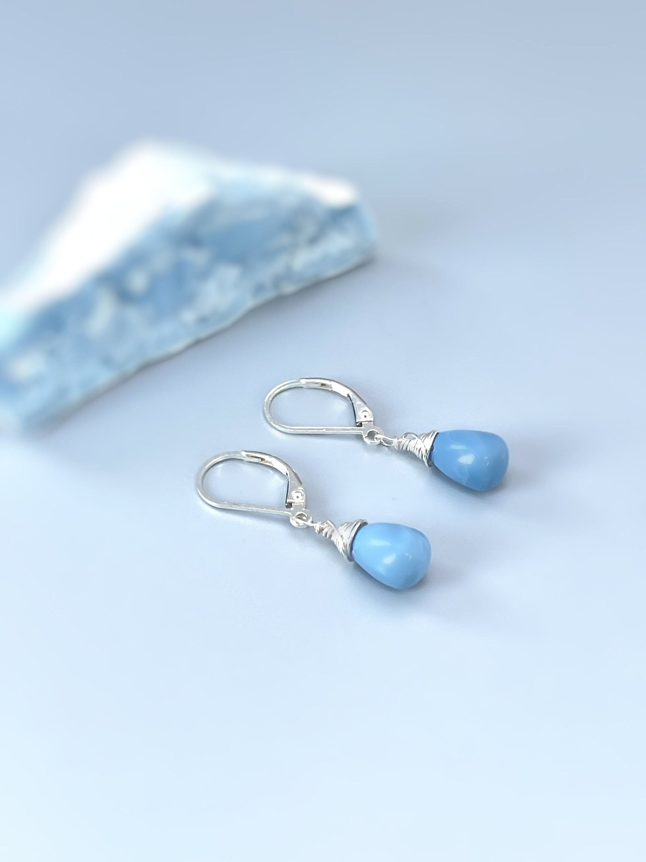 May include: A pair of silver teardrop earrings with light blue gemstone drops. The earrings have a simple design and are perfect for everyday wear.