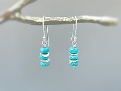 May include: Silver dangle earrings with turquoise blue beads and silver spacers. The earrings are hanging from a branch.