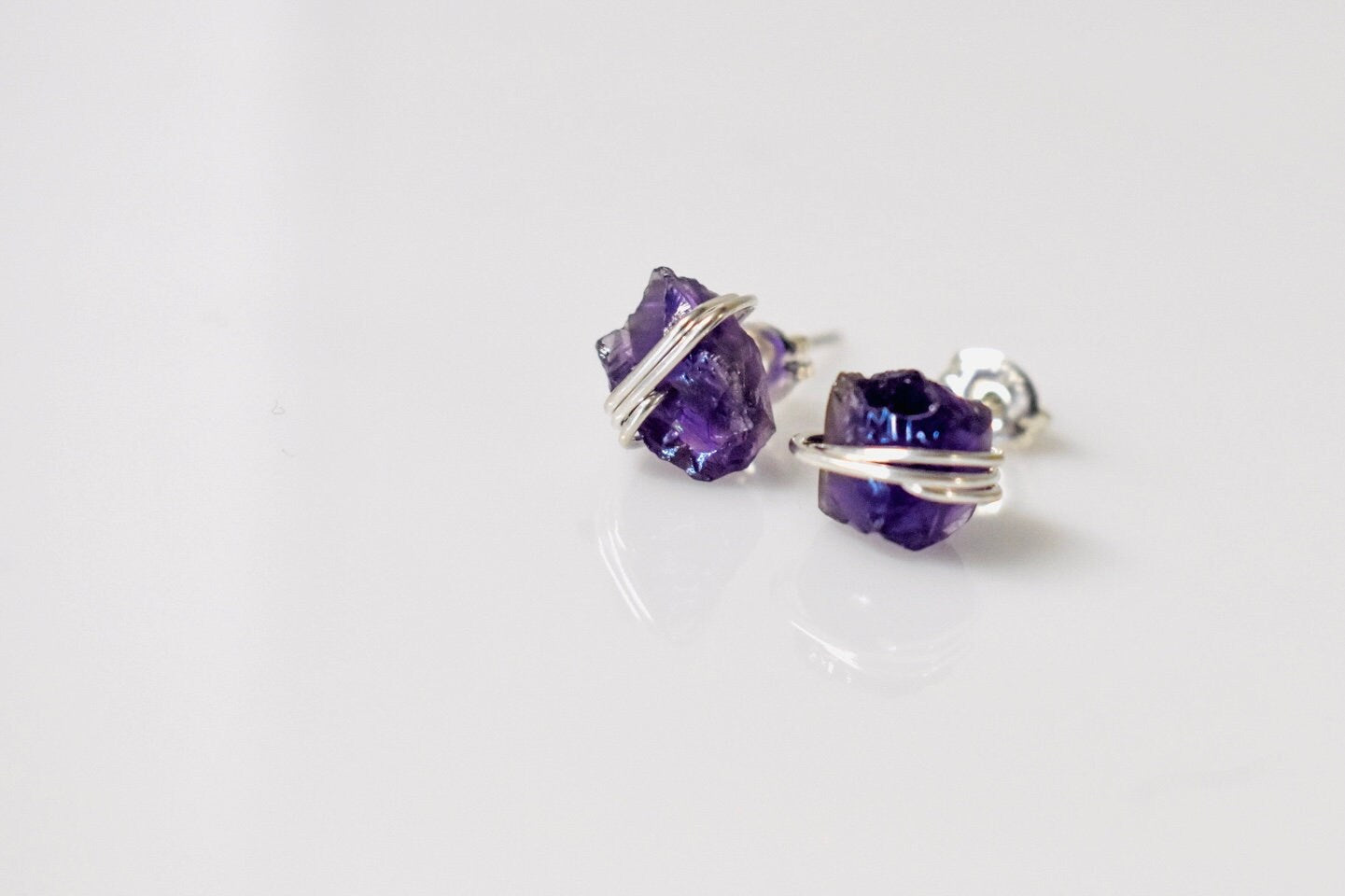 May include: A pair of silver wire wrapped amethyst earrings. The amethyst stones are a deep purple color and are wrapped in silver wire. The earrings are on a white background.