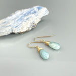May include: A pair of gold wire wrapped teardrop aquamarine earrings. The earrings are a light blue color and have a delicate, natural look.