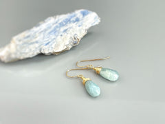 May include: A pair of gold wire wrapped teardrop aquamarine earrings. The earrings are a light blue color and have a delicate, natural look.