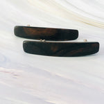 May include: Two dark brown wooden hair clips.  Each clip is rectangular with slightly rounded edges and features a subtle wood grain pattern. The clips are simple and elegant, showcasing the natural beauty of the wood. They appear to be made of a dark, polished wood, possibly ebony or a similar dark hardwood. The clips are positioned on a light gray marble-like surface.