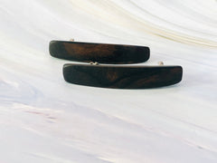 May include: Two dark brown wooden hair clips.  Each clip is rectangular with slightly rounded edges and features a subtle wood grain pattern. The clips are simple and elegant, showcasing the natural beauty of the wood. They appear to be made of a dark, polished wood, possibly ebony or a similar dark hardwood. The clips are positioned on a light gray marble-like surface.