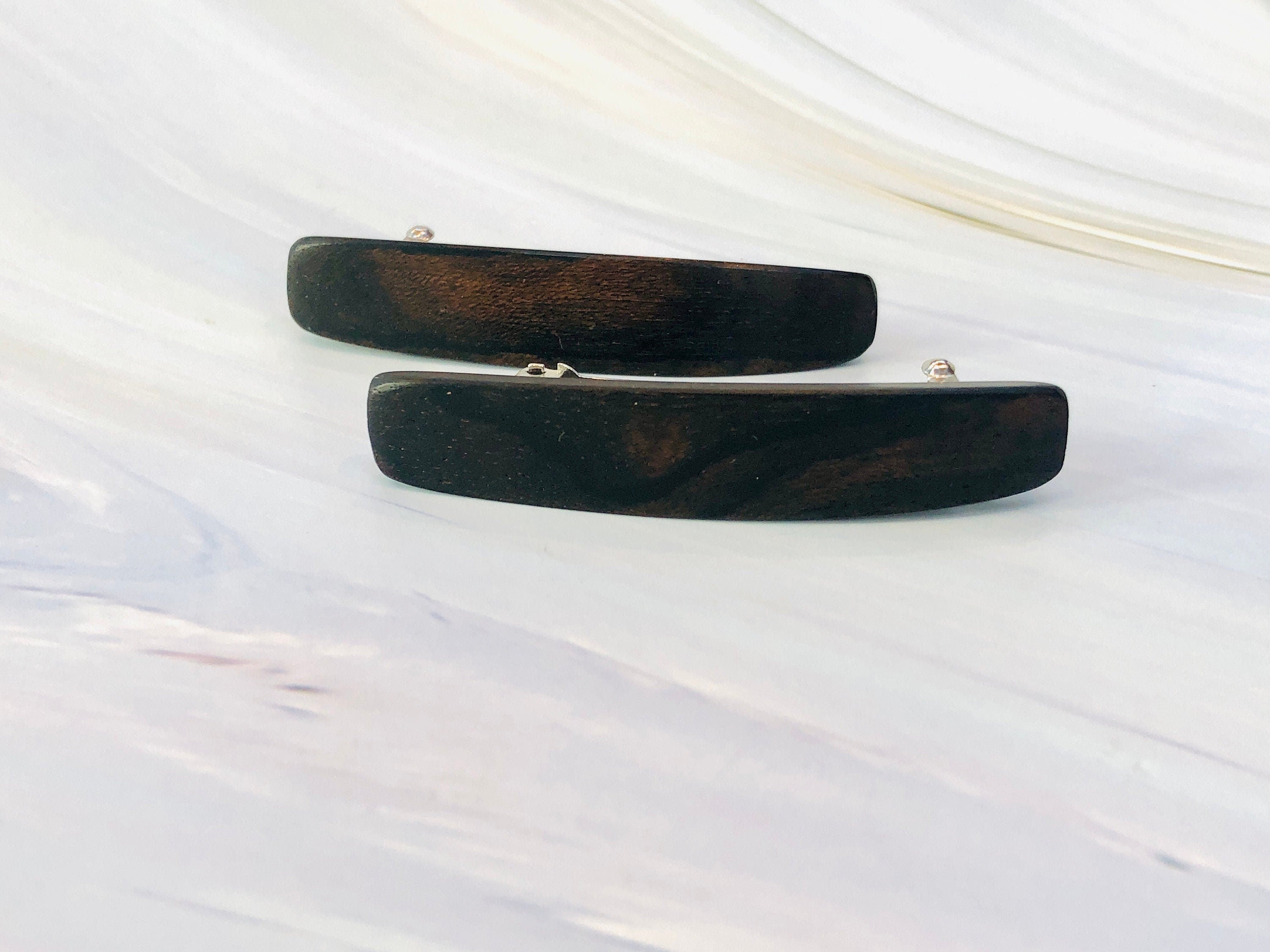 May include: Two dark brown wooden hair clips.  Each clip is rectangular with slightly rounded edges and features a subtle wood grain pattern. The clips are simple and elegant, showcasing the natural beauty of the wood. They appear to be made of a dark, polished wood, possibly ebony or a similar dark hardwood. The clips are positioned on a light gray marble-like surface.