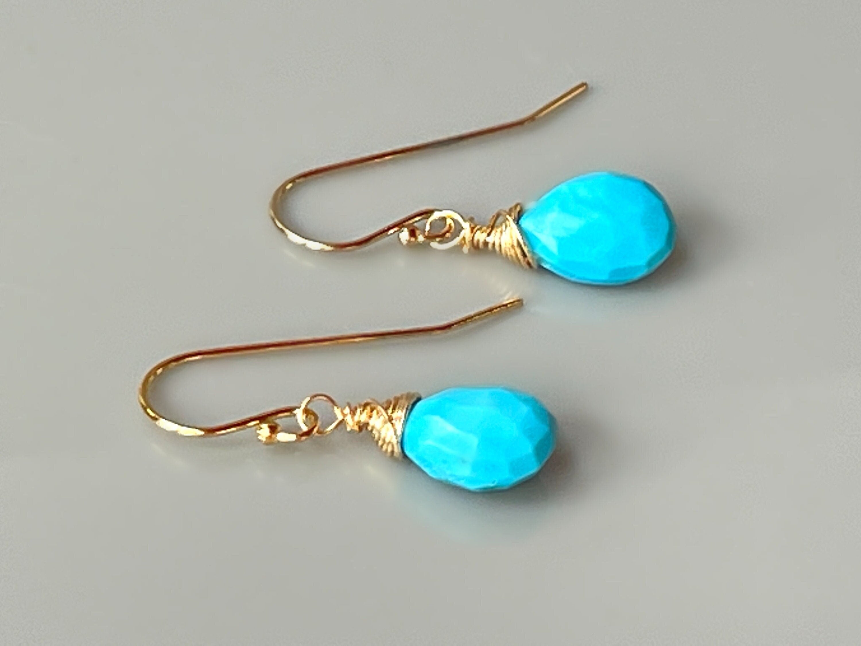May include: A pair of gold earrings with turquoise teardrop shaped stones. The stones are faceted and have a bright blue color. The earrings are simple and elegant.