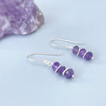 May include: A pair of silver dangle earrings with three faceted amethyst gemstones on each earring.
