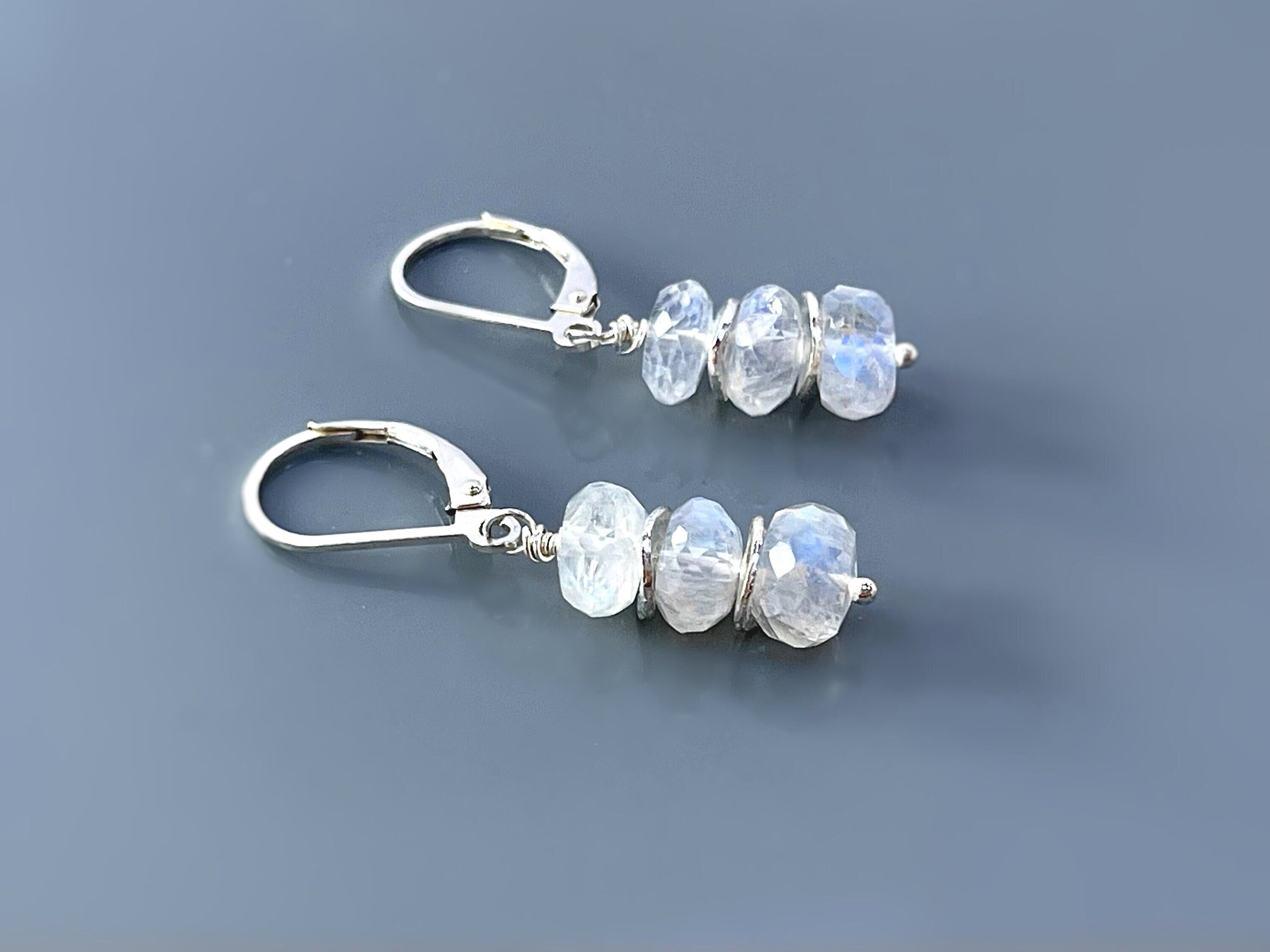 May include: Silver dangle earrings with three faceted moonstone beads each. The moonstone beads are a light white color with a slight iridescent sheen.