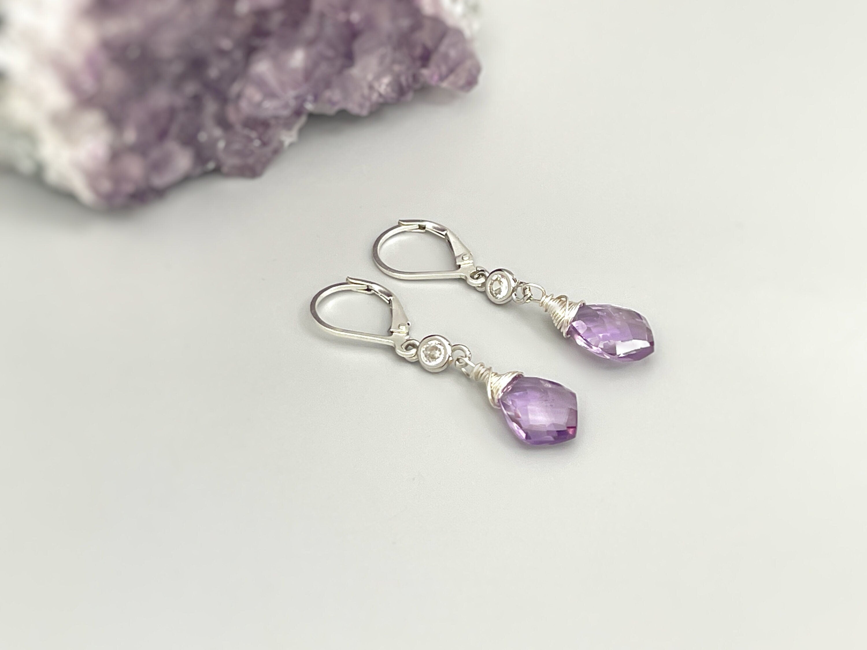 May include: A pair of silver earrings with amethyst gemstones. The earrings have a leverback closure and a small silver bead above the gemstone. The amethyst gemstones are a light purple color and have a faceted cut.