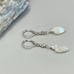 May include: A pair of silver dangle earrings with a white moonstone leaf charm. The earrings have a silver lever back closure and a small cluster of clear stones on the wire above the charm.