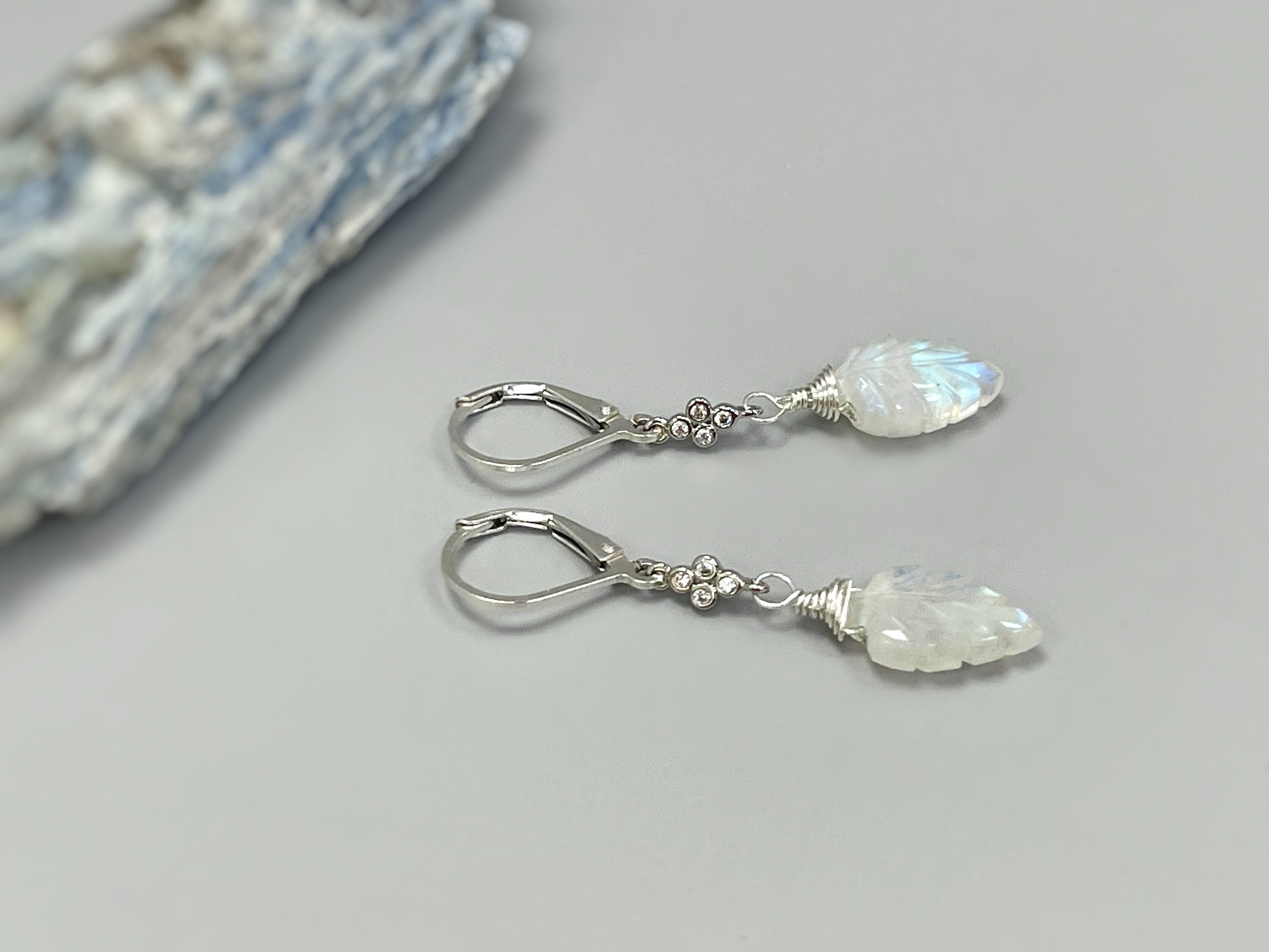 May include: A pair of silver dangle earrings with a white moonstone leaf charm. The earrings have a silver lever back closure and a small cluster of clear stones on the wire above the charm.