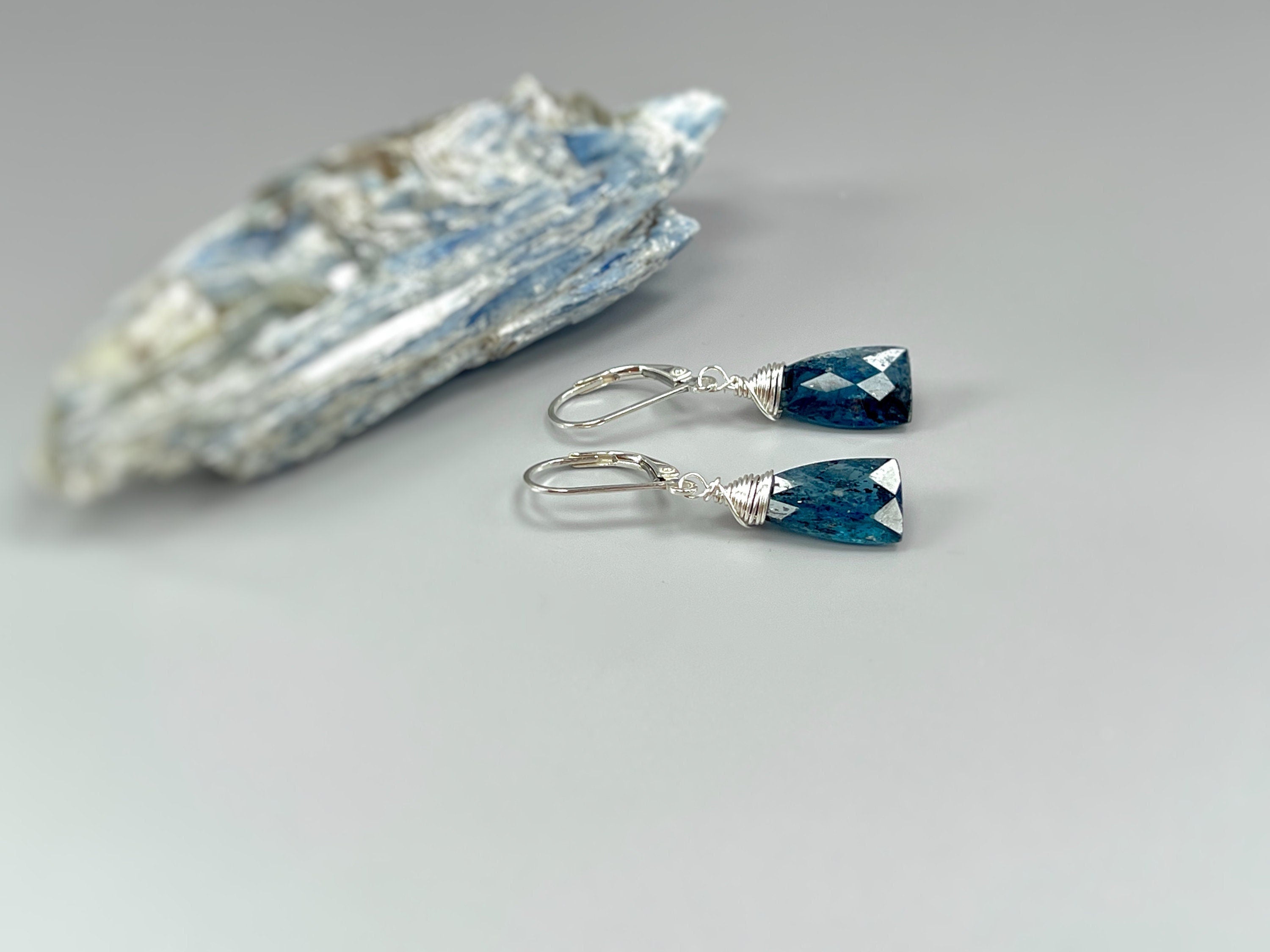 May include: A pair of silver earrings with blue gemstone drops. The gemstones are faceted and have a dark blue color. The earrings are hanging from silver hooks.
