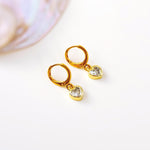 May include: A pair of gold-colored hoop earrings with heart-shaped pendants. Each heart pendant is encrusted with a clear, sparkling stone. The earrings are displayed on a white surface, with a blurred, iridescent background.