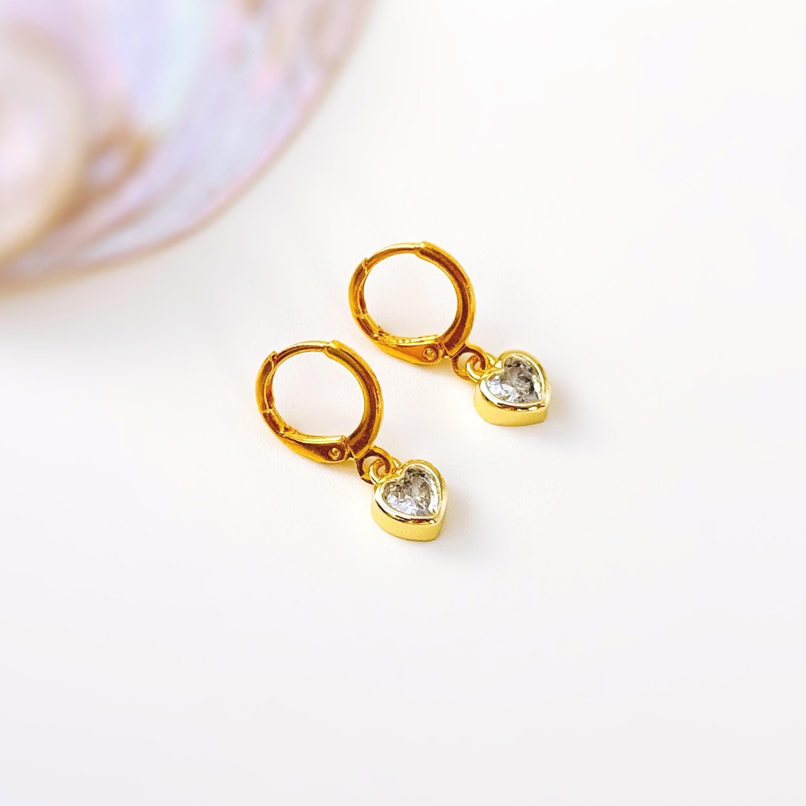May include: A pair of gold-colored hoop earrings with heart-shaped pendants. Each heart pendant is encrusted with a clear, sparkling stone. The earrings are displayed on a white surface, with a blurred, iridescent background.