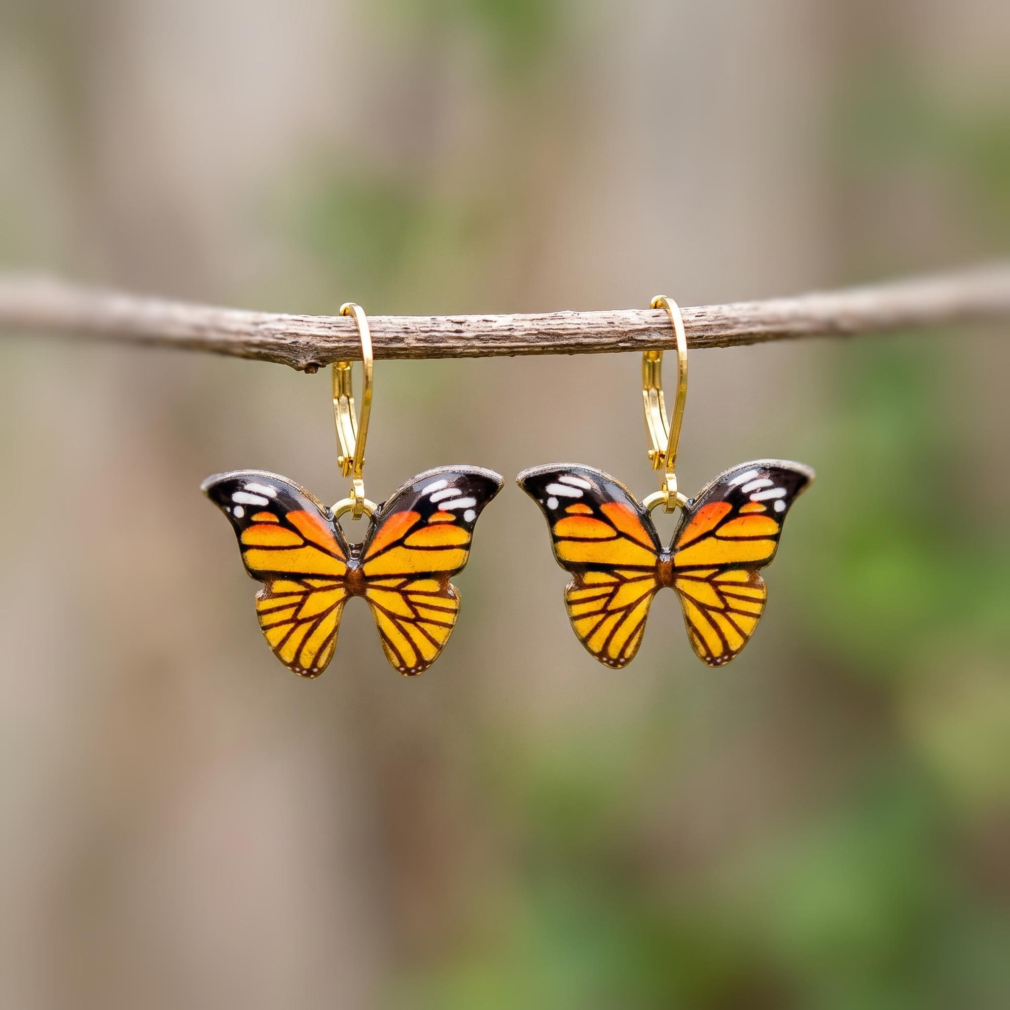 May include: A pair of dangle earrings featuring monarch butterfly charms with orange, black, and white wing patterns. The butterflies are suspended from gold-toned metal lever-back hooks, showcasing detailed wing veins and symmetrical markings.