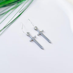 May include: A pair of silver-colored sword-shaped earrings with a hook closure. The earrings are displayed on a white surface with a few green leaves in the background. The swords have a detailed hilt and blade.