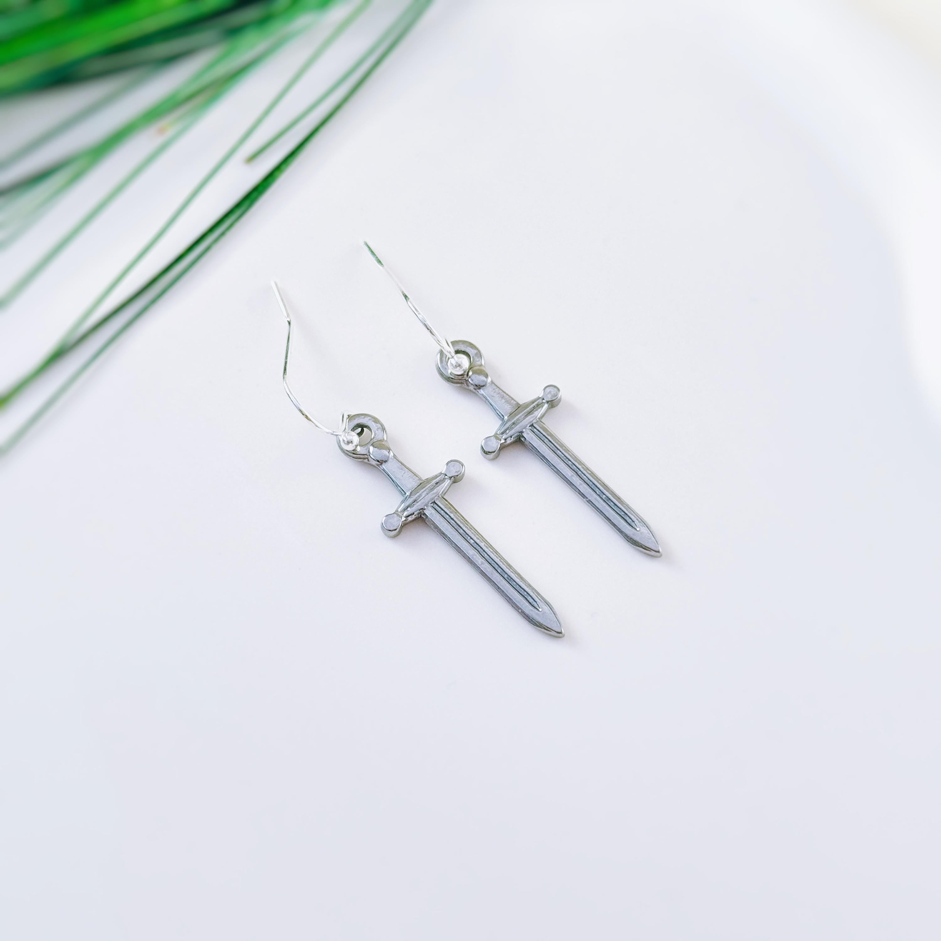 May include: A pair of silver-colored sword-shaped earrings with a hook closure. The earrings are displayed on a white surface with a few green leaves in the background. The swords have a detailed hilt and blade.
