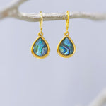 May include: A pair of gold hoop earrings featuring teardrop-shaped pendants with iridescent abalone shell inlays. The earrings have a polished gold finish and a hinged closure mechanism, showcasing vibrant blue and green natural shell patterns.