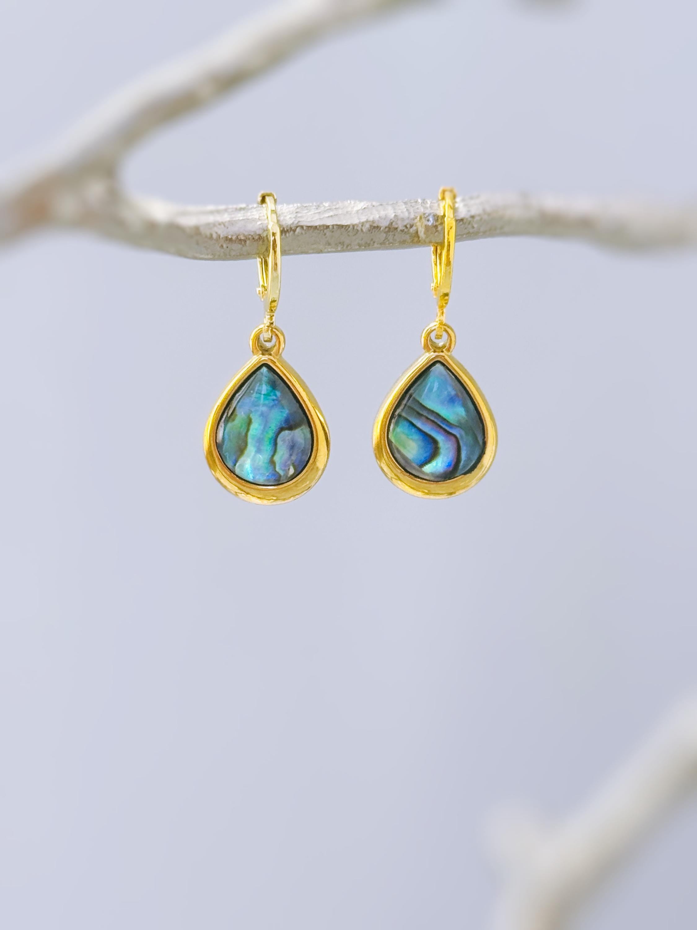 May include: A pair of gold hoop earrings featuring teardrop-shaped pendants with iridescent abalone shell inlays. The earrings have a polished gold finish and a hinged closure mechanism, showcasing vibrant blue and green natural shell patterns.
