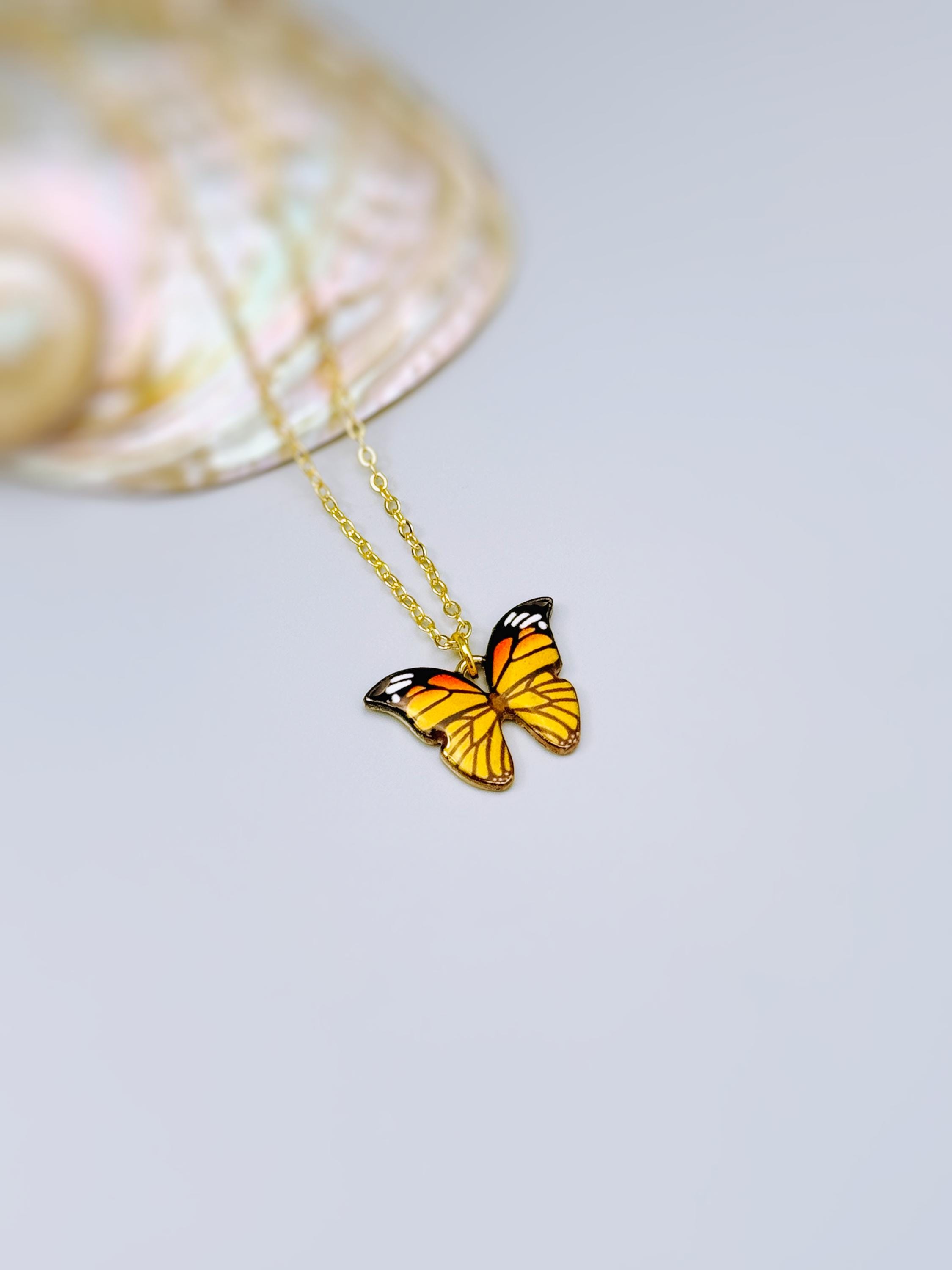 May include: A gold-toned chain necklace featuring a butterfly pendant with orange, yellow, black, and white enamel detailing. The butterfly wings are symmetrically patterned with black veins and white spots along the outer edges of the wings.