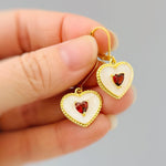 May include: Gold-tone heart-shaped earrings with a white enamel border and a small red heart-shaped stone in the center. The earrings have a lever-back closure. The earrings are held in a hand.