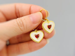 May include: Gold-tone heart-shaped earrings with a white enamel border and a small red heart-shaped stone in the center. The earrings have a lever-back closure. The earrings are held in a hand.