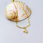 May include: A gold-colored necklace with a small bunny pendant. The necklace is made of a chain link design. The pendant is a small bunny with a textured surface. The necklace is resting on a large, iridescent seashell.
