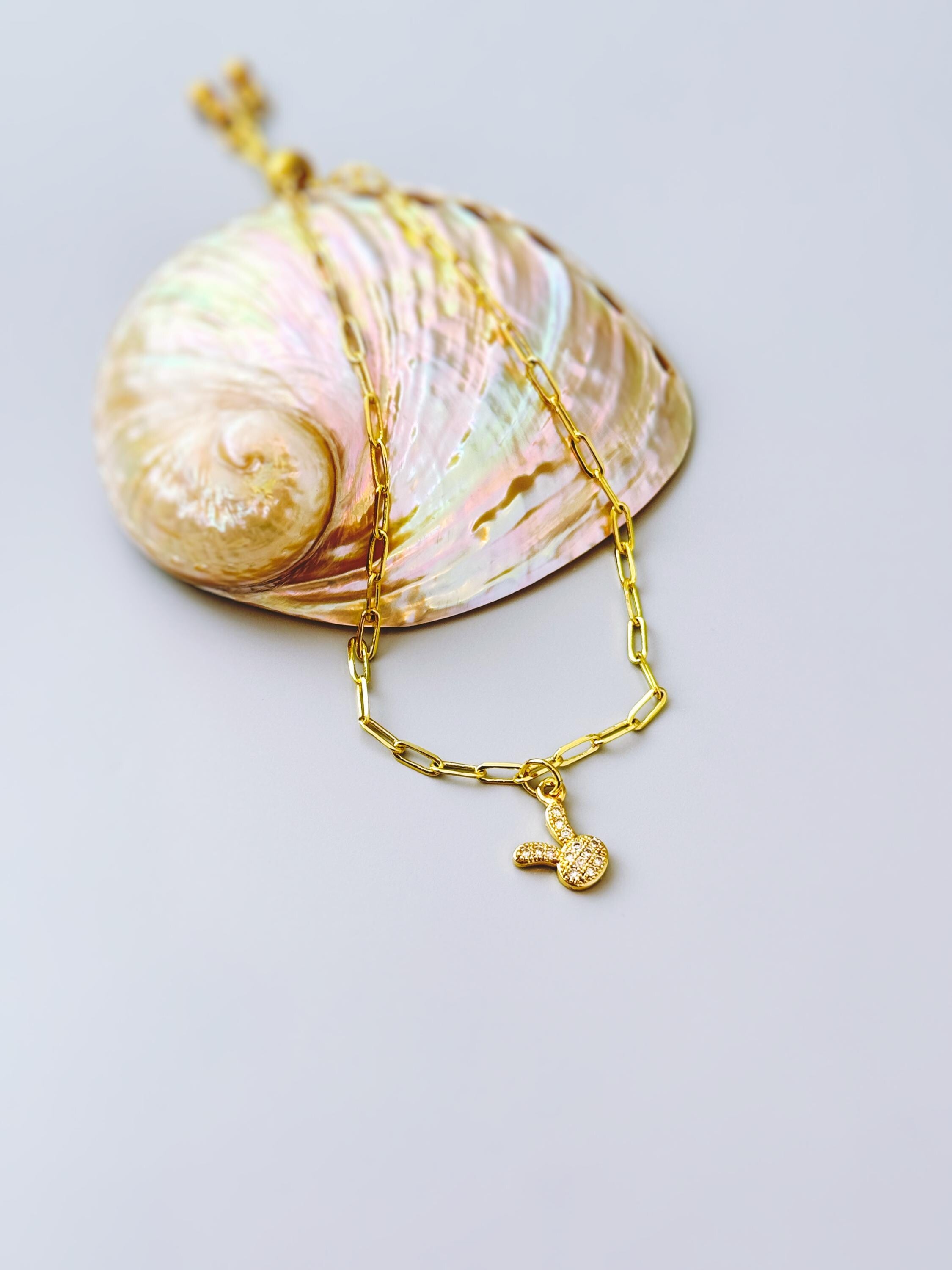 May include: A gold-colored necklace with a small bunny pendant. The necklace is made of a chain link design. The pendant is a small bunny with a textured surface. The necklace is resting on a large, iridescent seashell.